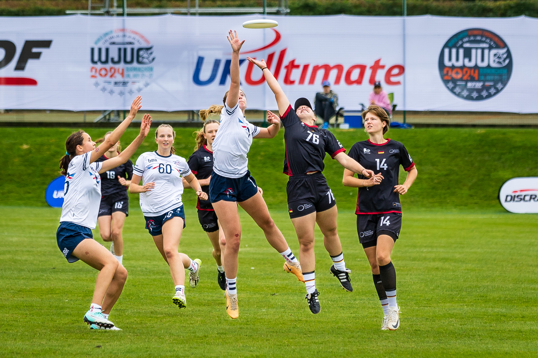 Athletes compete for a disc in the air above them, on a field in front of the WJUC tournament logo signage