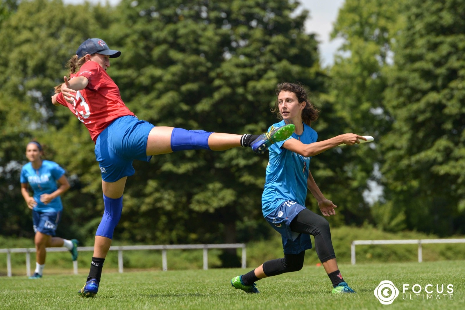 A woman in a blue ITALIA jersey looks to throw the disc past a player in a red jersey attempting a foot block.
