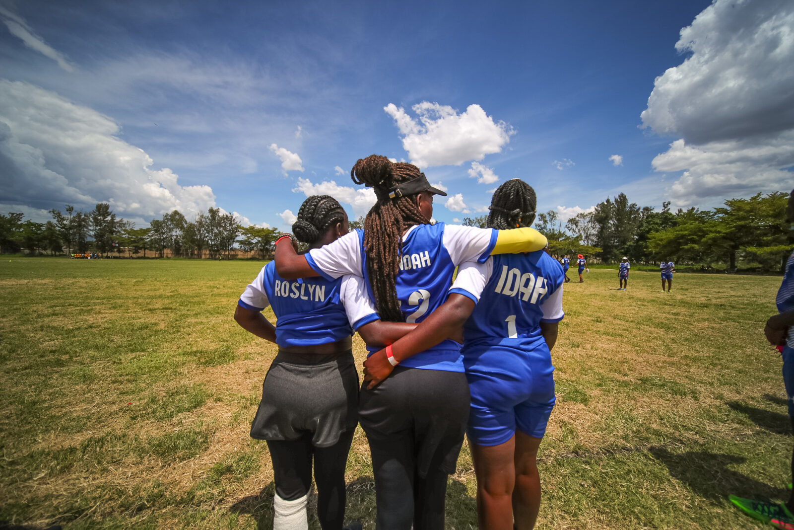 Three women wearing blue jerseys stand with their arms around each other and backs to the camera, looking out over a frisbee field.