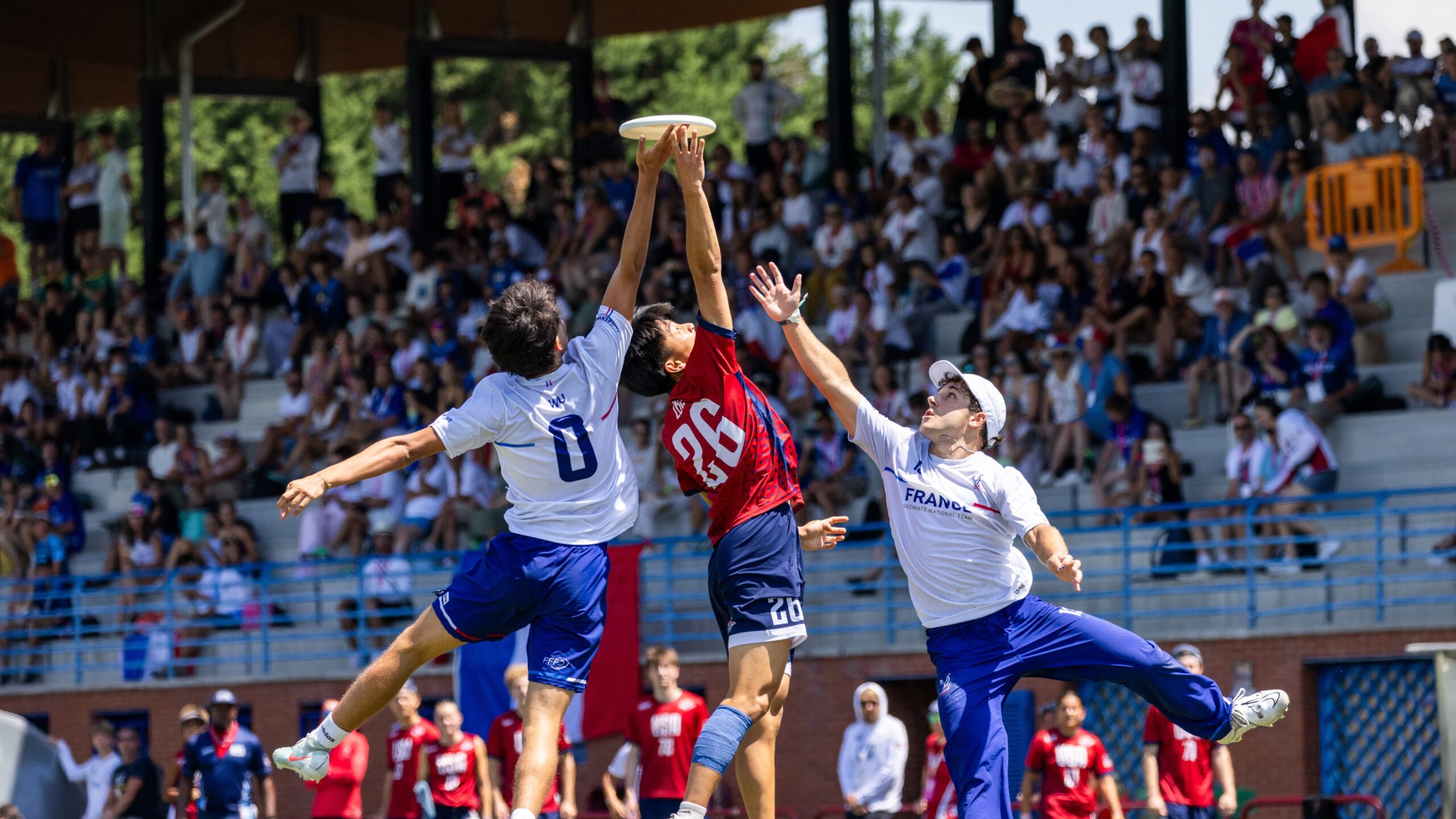 Three junior ultimate frisbee players jump to catch the disc during the WJUC 2026 game, with spectators in the background.