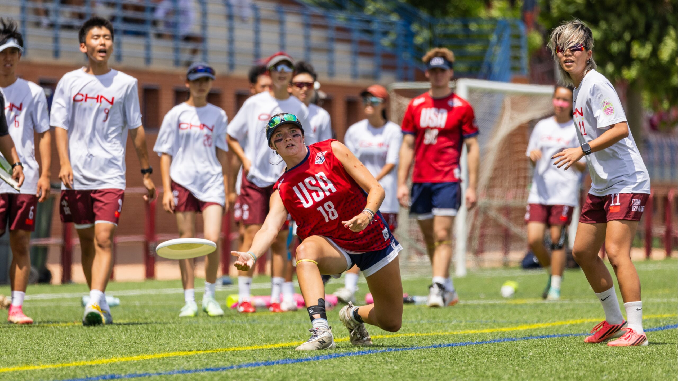 A USA player stretches low to release the disc during a mixed ultimate frisbee game, while players from China and the USA watch the play from the background on a sunny outdoor field.