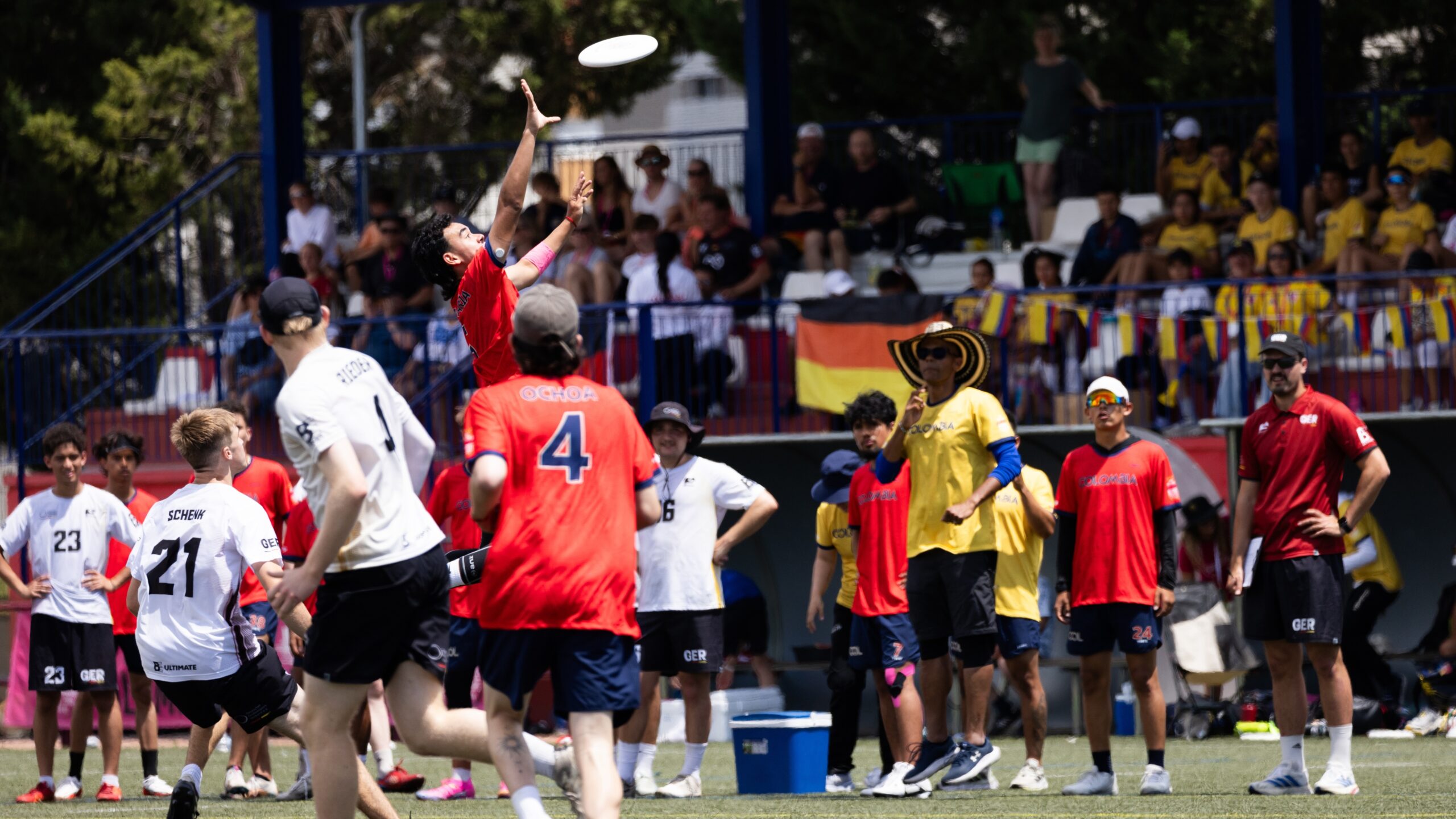 A player in a red jersey leaps high to catch a flying disc during an outdoor ultimate frisbee match, while players in white and red jerseys watch the play. Team staff and substitutes stand along the sideline, with spectators and national flags visible in the background under bright sunny conditions.