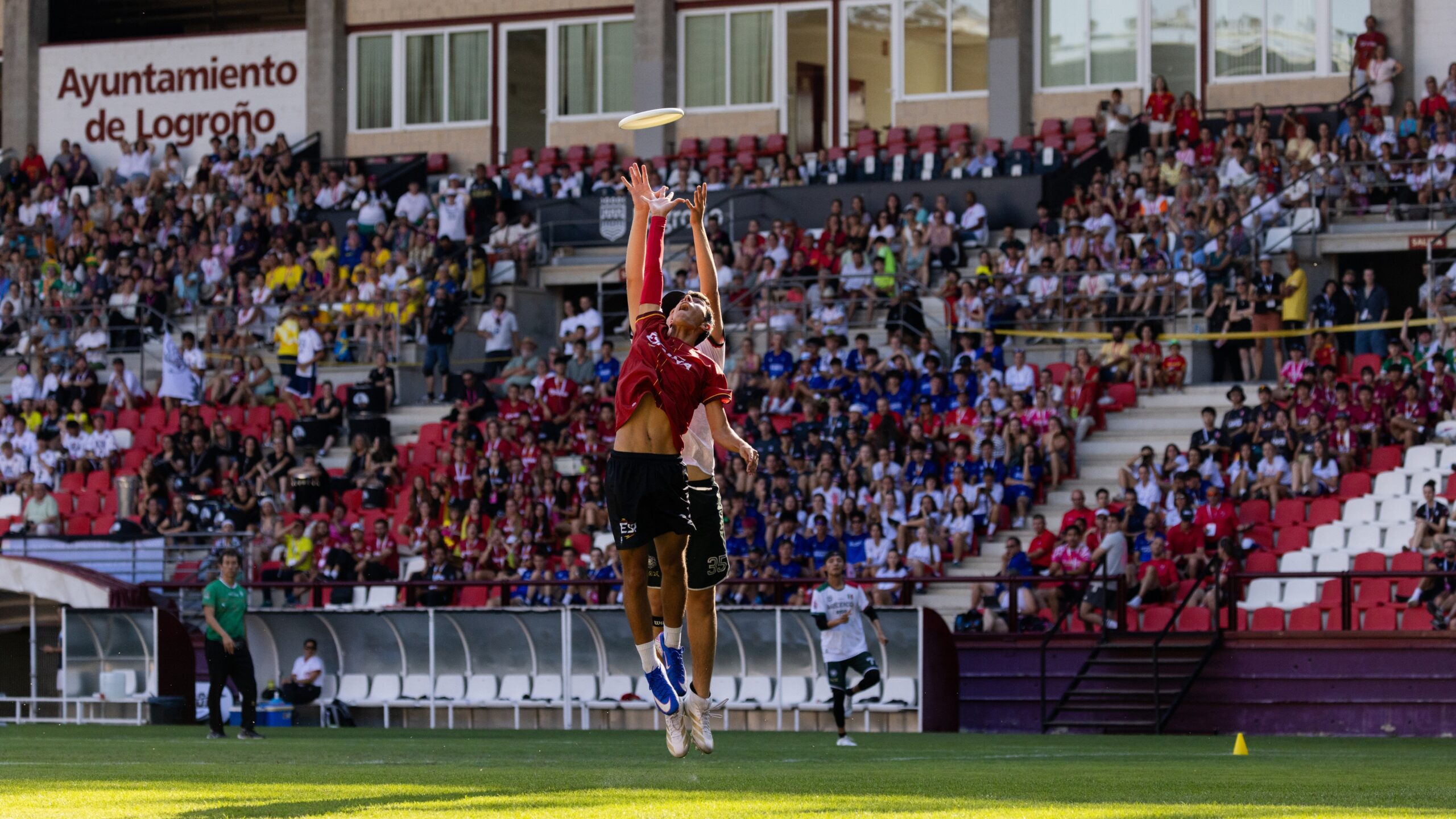Two players leap for the disc during the opening day of the 2026 World Junior Ultimate Championships in front of spectators at the stadium.