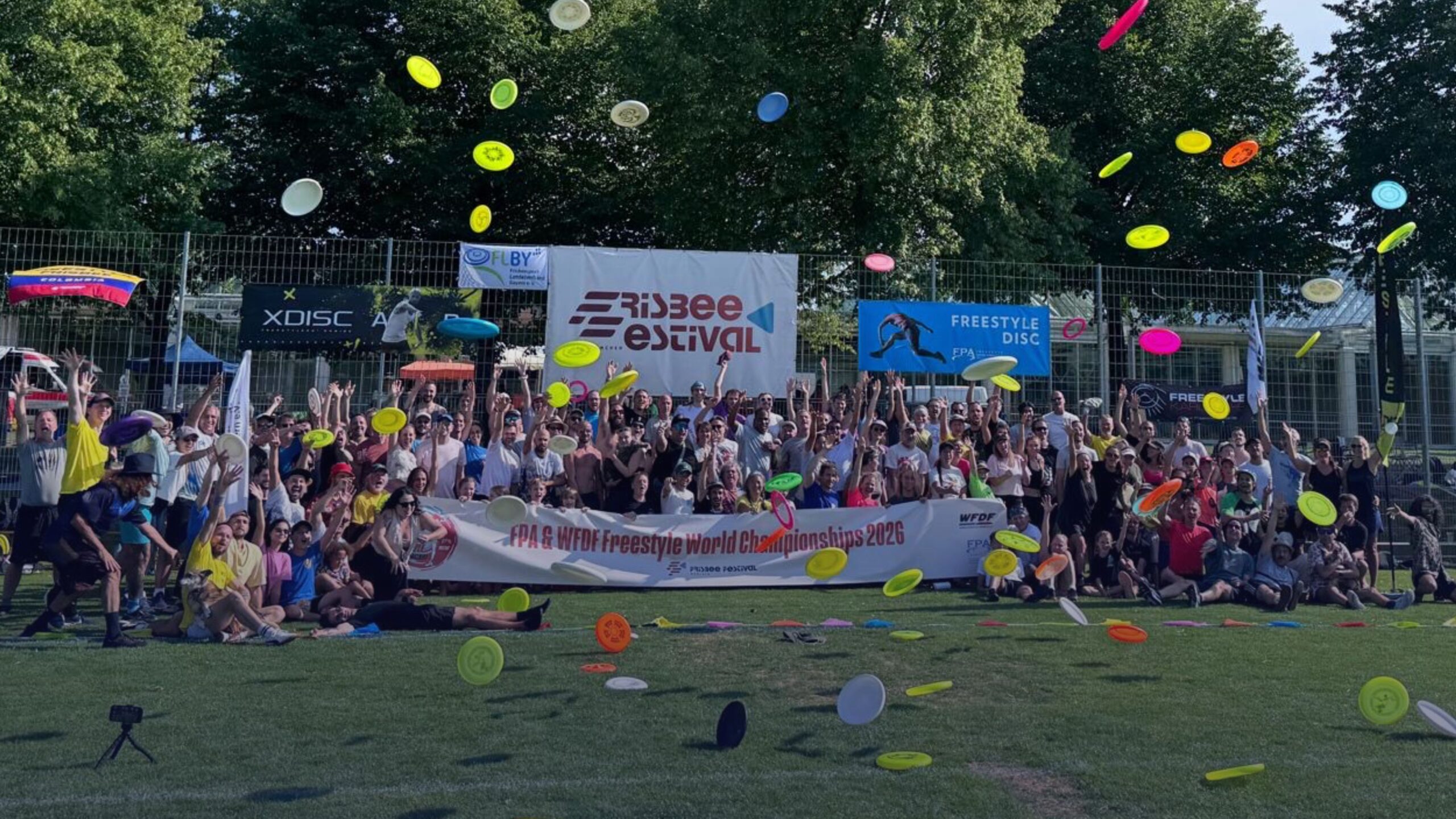 Group of athletes and spectators celebrating at the FPA & WFDF Freestyle World Championships 2026, with colorful flying discs filling the air and the Freestyle Disc Festival backdrop visible behind them.