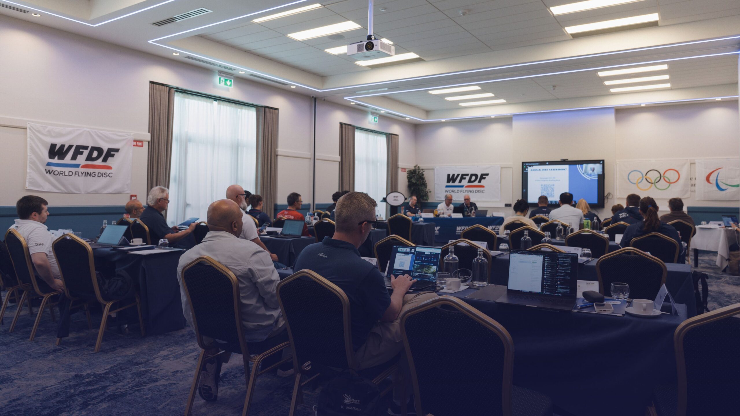 WFDF Annual Congress 2026 meeting in a conference room, with attendees seated at tables facing a presentation screen and WFDF banners, with Olympic and Paralympic symbols visible on the wall.