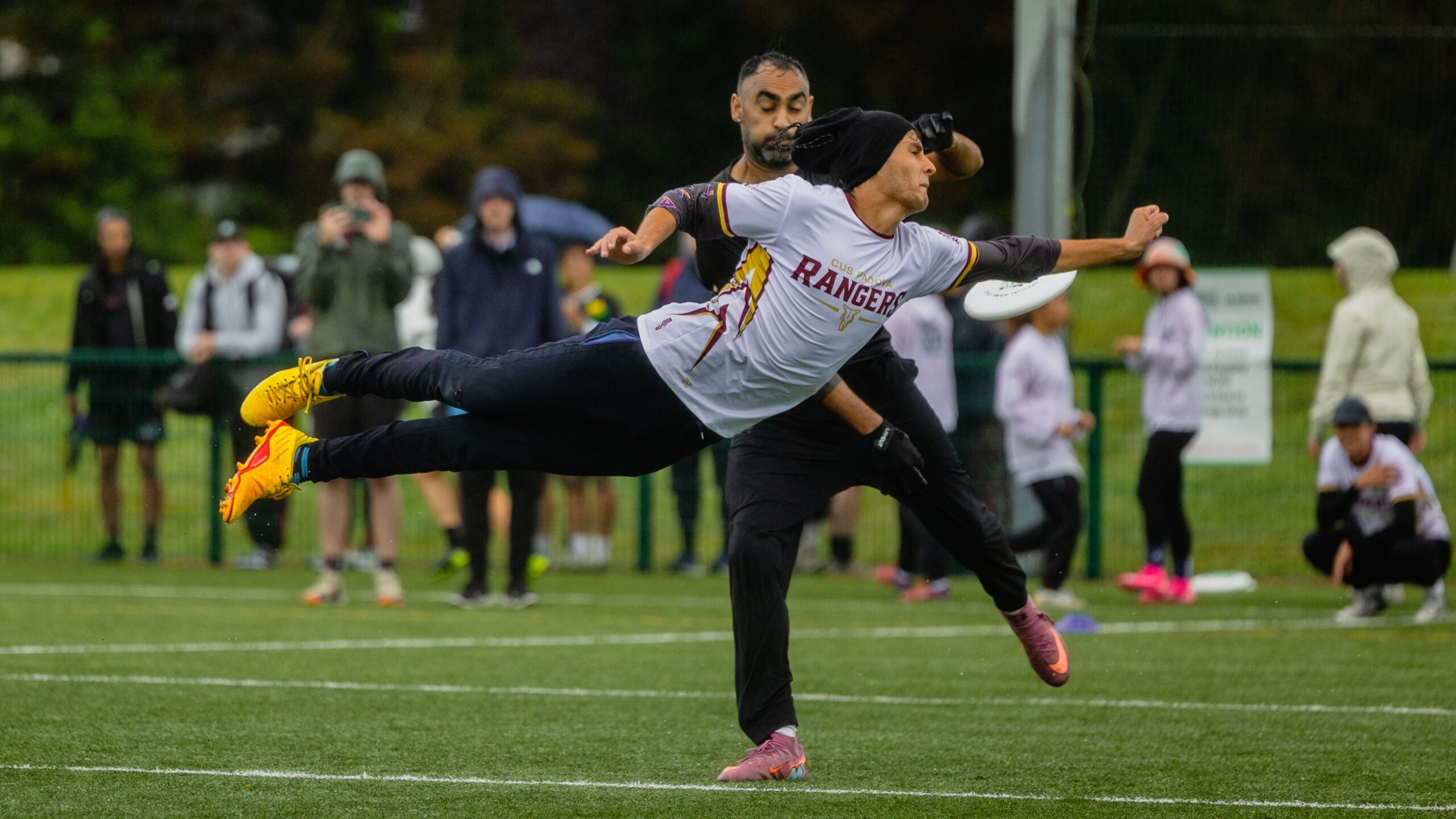An ultimate frisbee game on a wet field, with a player in a white “Rangers” jersey diving horizontally to catch or throw the disc, while another player and spectators stand in the background