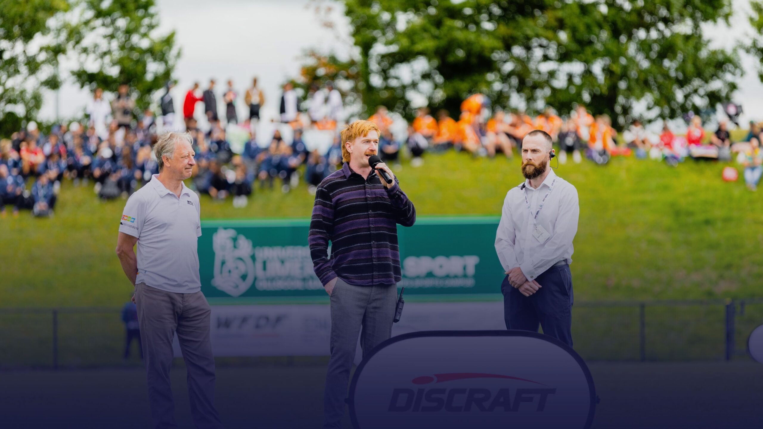 Opening ceremony of the WFDF 2026 World Ultimate Club Championships in Limerick, with a speaker addressing the crowd on the field alongside two event officials, as spectators watch from the grassy hillside.
